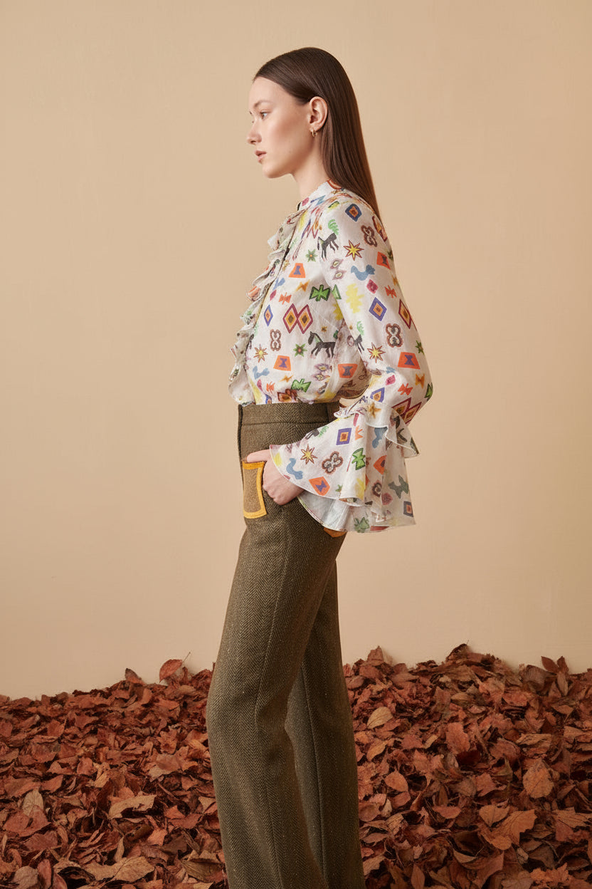 Woman wearing a colorful patterned blouse and brown pants with yellow accents, standing on a beige background.