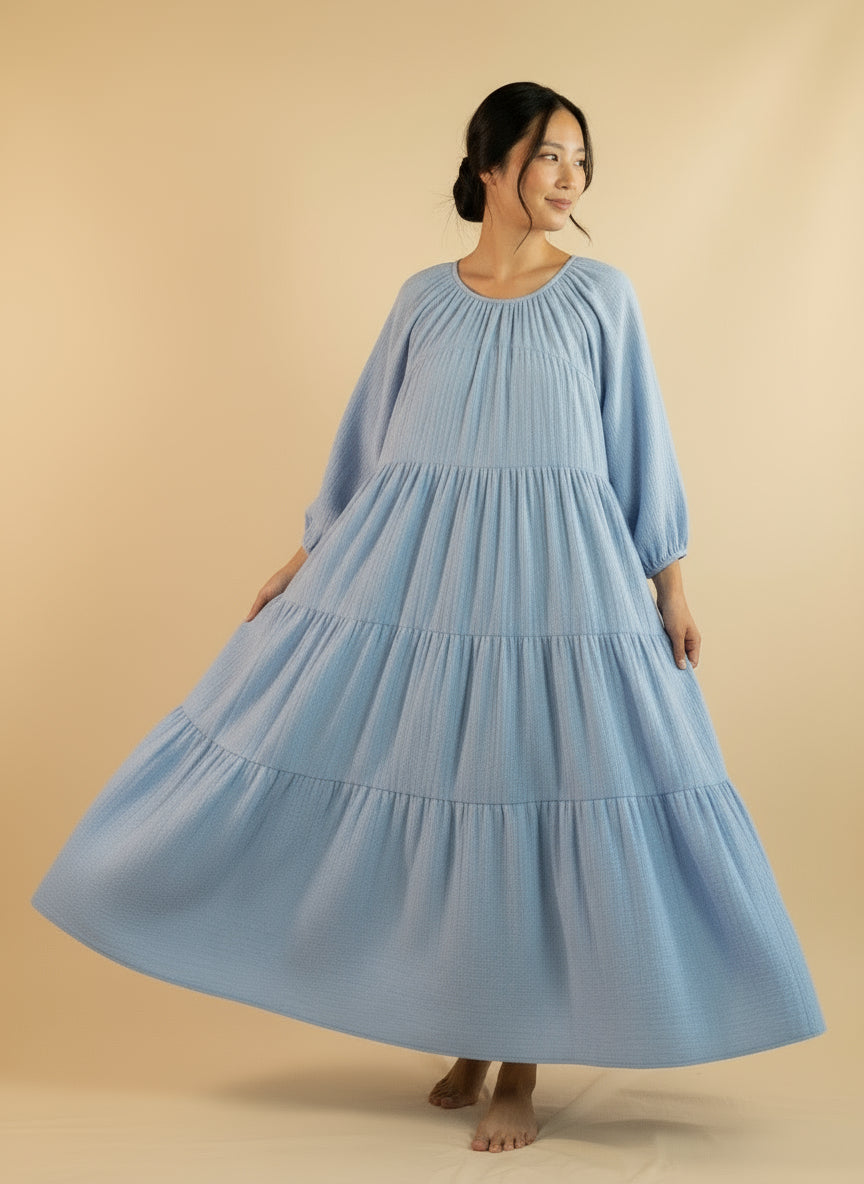 A woman modeling a blue maxi dress with a gathered waist and ruffled detailing, posing against a beige background.