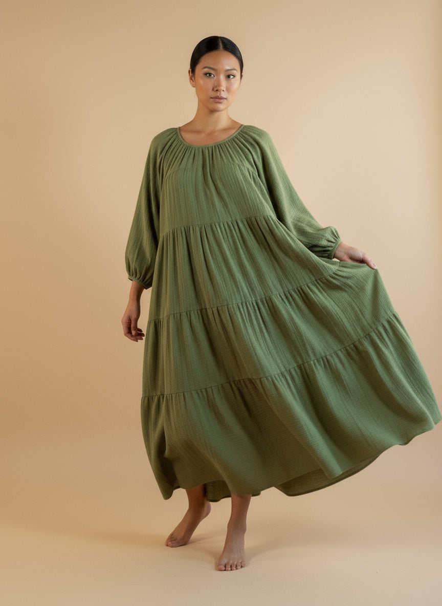 A woman standing on an orange background wearing a green, solid-colored maxi dress with a flowing skirt and a round neckline.
