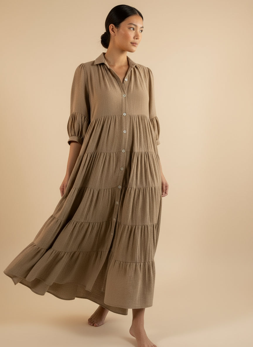 A woman standing and modeling a beige maxi sleeved dress with a V-neckline and pleated details.